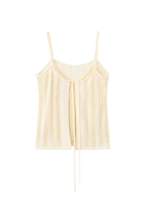 Femme portant un haut sans manches en maille jaune pâle et un pantalon large assorti, lunettes de soleil foncées, debout devant un fond blanc uni.