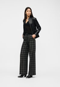 Γυναίκα με μακριά σκουρόχρωμα μαλλιά, φοράει μαύρο δερμάτινο μπουφάν, μαύρο τοπ, γκρι καρό φαρδιά παντελόνια και μαύρα μποτάκια αστραγάλου με ασημί άκρες.