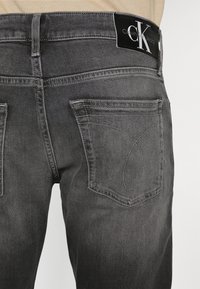 Ciemnoszare dżinsy z denimu o dopasowanym kroju, wyposażone w pas z logo Calvin Klein oraz charakterystyczne przeszycia na tylnej kieszeni.