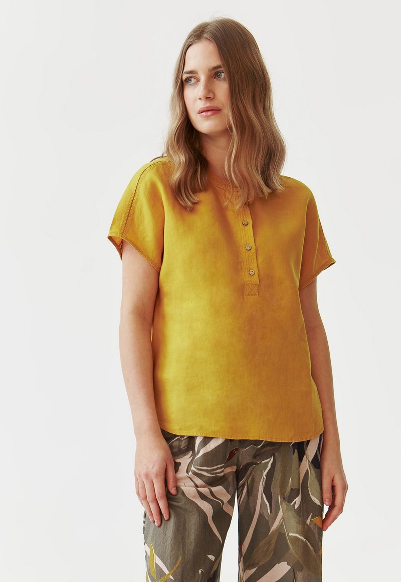Camicia di lino gialla con maniche corte, dotata di chiusura a bottoni sul collo e tessuto a trama. Abbinata a pantaloni verdi a fantasia.