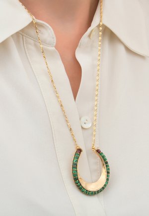 Un collier en or présente un pendentif courbé incrusté d'accents verts et violets, suspendu à une chaîne texturée contre une chemise claire.