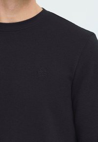 Črna teksturirana srajca s krožnim izrezom z diskretnim embosiranim kubičnim logotipom na levi strani prsnega koša, izdelana iz mehkega in zračnega materiala.