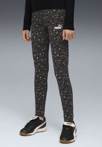Persona vestita con leggings Puma leopardati neri e grigi e sneakers Puma nere, in piedi su una superficie grigia contro uno sfondo grigio.