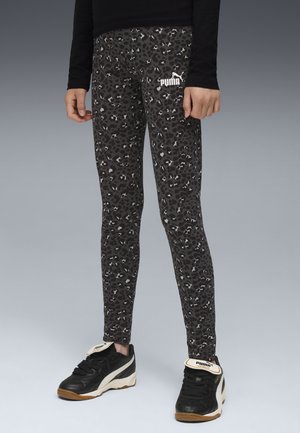 Persona vestita con leggings Puma leopardati neri e grigi e sneakers Puma nere, in piedi su una superficie grigia contro uno sfondo grigio.