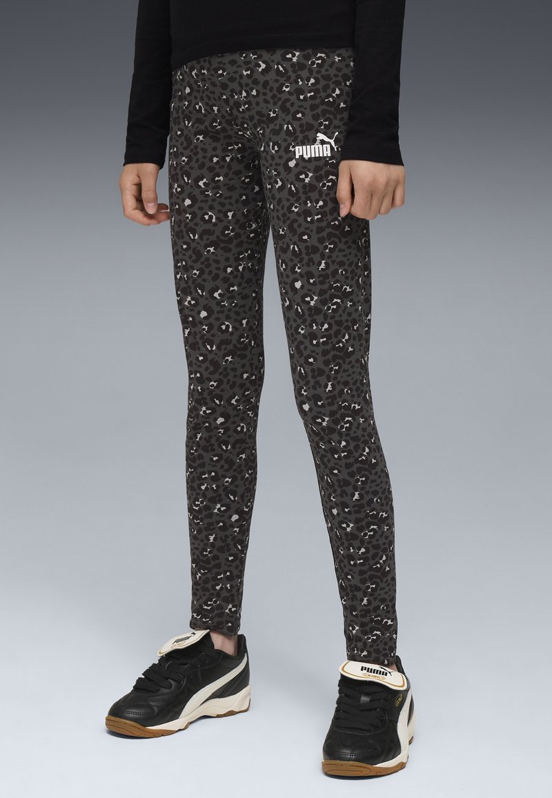 Persona vestita con leggings Puma leopardati neri e grigi e sneakers Puma nere, in piedi su una superficie grigia contro uno sfondo grigio.