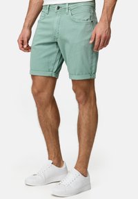 Helle grüne Denim-Shorts mit umgeschlagenen Säumen, im Fünf-Taschen-Design und einem Frontknopfverschluss. Kombiniert mit weißen Low-Top-Sneakern.