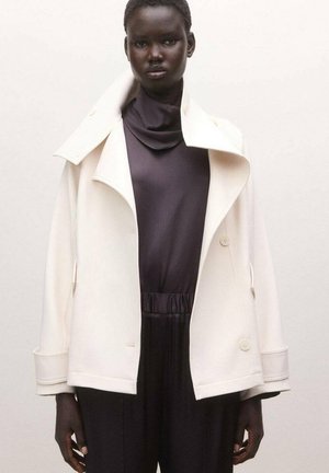 Mujer con chaqueta blanca de cuello alto sobre una blusa negra y pantalones negros, de pie frente a un fondo claro y liso.