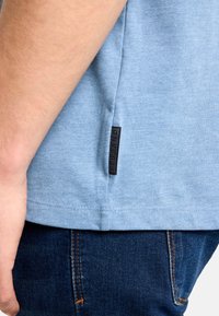 Svetlomodré bavlnené tričko s krátkymi rukávmi a bočnou značkou, v kombinácii s tmavými džínsami z denimu. Viditeľný šev lemovaný pozdĺž okraja.