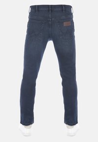 Vue arrière d'un jean en denim bleu foncé porté avec des baskets blanches, montrant deux poches arrière et une étiquette en cuir sur la poche droite.