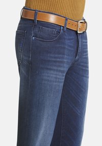 Un jean en denim bleu foncé présente un design droit avec un effet de décoloration visible, accompagné d'une ceinture en cuir marron. Des poches et des détails de couture sont présents.