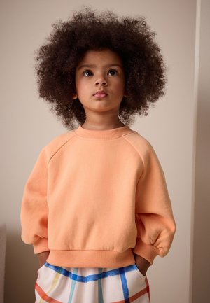 Mladé dieťa s kučeravými vlasmi, oblečené v broskyñovej mikine a farebných kockovaných nohaviciach, pozerá mierne nahor so založenými rukami v džepoch.