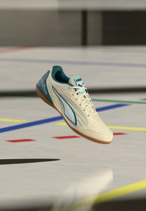 Chaussure de sport d’intérieur Puma beige et bleu sarcelle flottant au-dessus d’un sol de gymnase avec des marquages colorés.