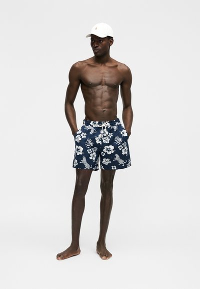 Särkideta mees, kes kannab valget nokamütsi ja tumesiniseid ujumispükse valgete lille- ja leopardimustritega, seisab käed taskutes.