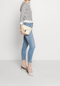 Cardigan de patrón cremoso, jeans ajustados de color azul claro, sandalias de tacón en blanco y negro, sosteniendo un clutch amarillo con solapa curva y detalle metálico.
