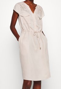 Robe beige à manches courtes avec un col en V, deux poches avant, une taille à cordon et des accents cloutés sur les épaules. Tissu doux et léger.