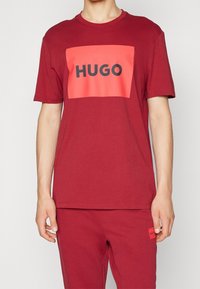 Camiseta de algodón roja con un gráfico rectangular rosa que presenta "HUGO" en letras negritas negras, cuello redondo y corte relajado.
