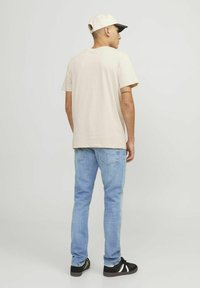 Camiseta de algodón beige, corte relajado, combinada con jeans de mezclilla azul claro, zapatillas negras y blancas, y una gorra color crema con visera negra.