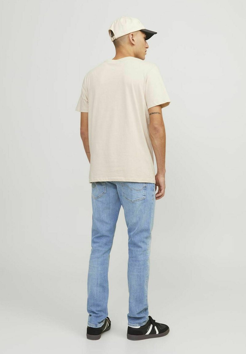Beige Baumwoll-T-Shirt, lockere Passform, getragen mit hellblauen Jeans, schwarz-weißen Sneakers und einer cremefarbenen Mütze mit schwarzem Schirm.