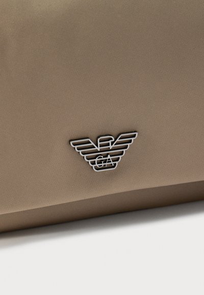 Κοντινό πλάνο μπεζ υφάσματος με ένα μαύρο και λευκό έμβλημα αετού Emporio Armani ραμμένο.