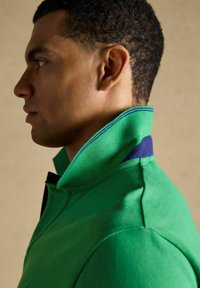 Homme de profil portant une chemise verte à col relevé, fond beige, cheveux courts et bouclés.