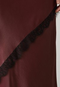 Main soulevant un tissu satin bordeaux foncé avec une bordure en dentelle noire le long du bord diagonal.