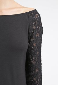 Černé tričko s přiléhavým střihem a dlouhými průhlednými krajkovými rukávy s květinovým vzorem. Výstřih je široký a odhaluje ramena.