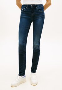 Jeans azul-escuros de corte justo feitos de ganga, com um design clássico de cinco bolsos e cintura com botões, combinados com ténis brancos.