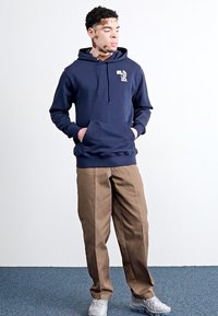 Sweat à capuche bleu marine avec poche kangourou, associé à un pantalon droit marron clair. Modèle portant des baskets grises.