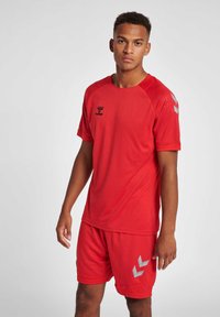 Rood sportoutfit bestaande uit een kortemouwen textuurtrui en bijpassende shorts. Kenmerkt zich door een zwart logo en zilveren chevronaccenten.