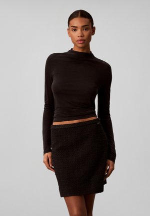 Femme aux cheveux attachés portant un haut noir ajusté à manches longues et une jupe noire texturée longueur genou, debout devant un fond uni.