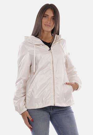 Donna con capelli castani lisci che indossa una giacca beige chiaro con cappuccio e cerniera e jeans blu, in piedi con una mano in tasca.