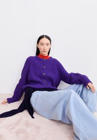 Lila gerippter Strickcardigan mit Rundhalsausschnitt und schwarzen Knöpfen, kombiniert mit hellblauen, lockeren Jeans und einem roten Rollkragenpullover darunter.