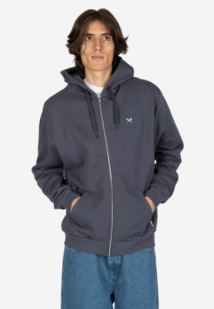 Junger Mann mit mittelbraunen Haaren, der eine dunkelgraue Kapuzenjacke mit Reißverschluss trägt, die Hände in den Taschen hat und blaue Jeans trägt, schaut leicht nach oben.