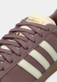 Ibolya színű bőr sportcipő krémszínű részletekkel, három fehér csíkkal és párnázott nyelvvel. Arany logó a nyelven, texturált fűzők.