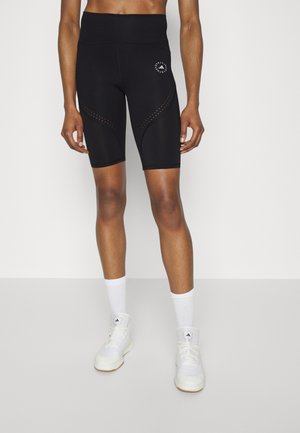 Person, die schwarze High-Waist-Radlerhosen mit perforierten Details sowie weiße Sportschuhe und Socken trägt, steht auf einem hellgrauen Boden.