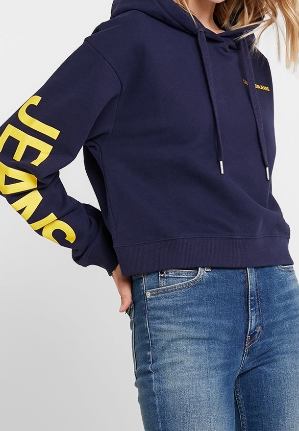 Kvinna som bär en marinblå kortärmad hoodie med gul "JEANS" text på ärmen, i kombination med blå denimjeans.