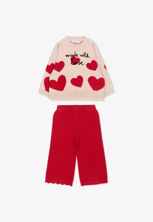 Pull beige à manches longues avec des cœurs rouges et le texte "made with love", porté avec un pantalon rouge à jambes larges et ourlets festonnés.