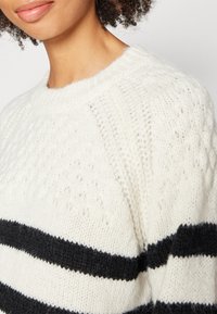 Primer plano de una persona que lleva un suéter de punto blanco con rayas horizontales negras y patrones texturizados en el hombro y el pecho.