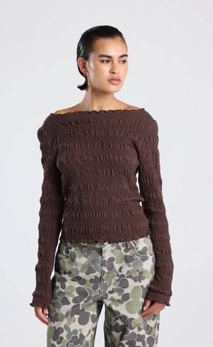Bruine off-the-shoulder top met textuur en geraffineerde stof; lange mouwen en subtiele ruffles langs de randen. Gecombineerd met broek met camouflagedessin.