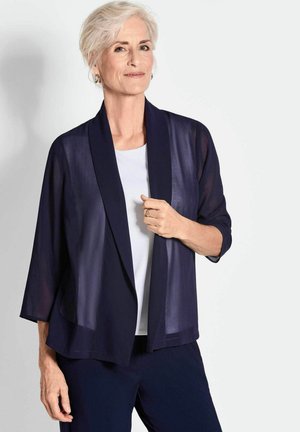 Cardigan trasparente blu navy con frontale drappeggiato, colletto a scialle e maniche a tre quarti, indossato sopra una maglietta bianca. Tessuto morbido e leggero.