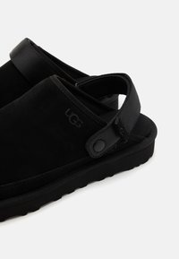 Black suede slip-on shoes with a textured strap and button accent. Features a thick sole for cushioning and support.