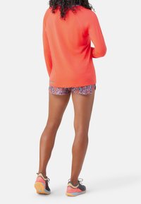 Dlouhý rukáv, jasně korálový sportovní top s raglánovými rukávy, doplněný květinově vzorovanými šortkami. Černé a růžové běžecké boty s texturovanou podrážkou.