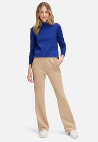 Blå tröja med hög krage och långa ärmar, ihop med beigefärgade utsvängda byxor. Utstyrseln inkluderar vita sneakers och har en slät textur.