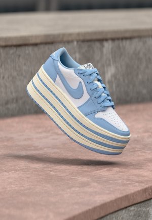 Baskets Nike blanches et bleu clair avec une semelle superposée surdimensionnée, flottant au-dessus d'une surface de pierre rosée et d'un fond en pierre grise.