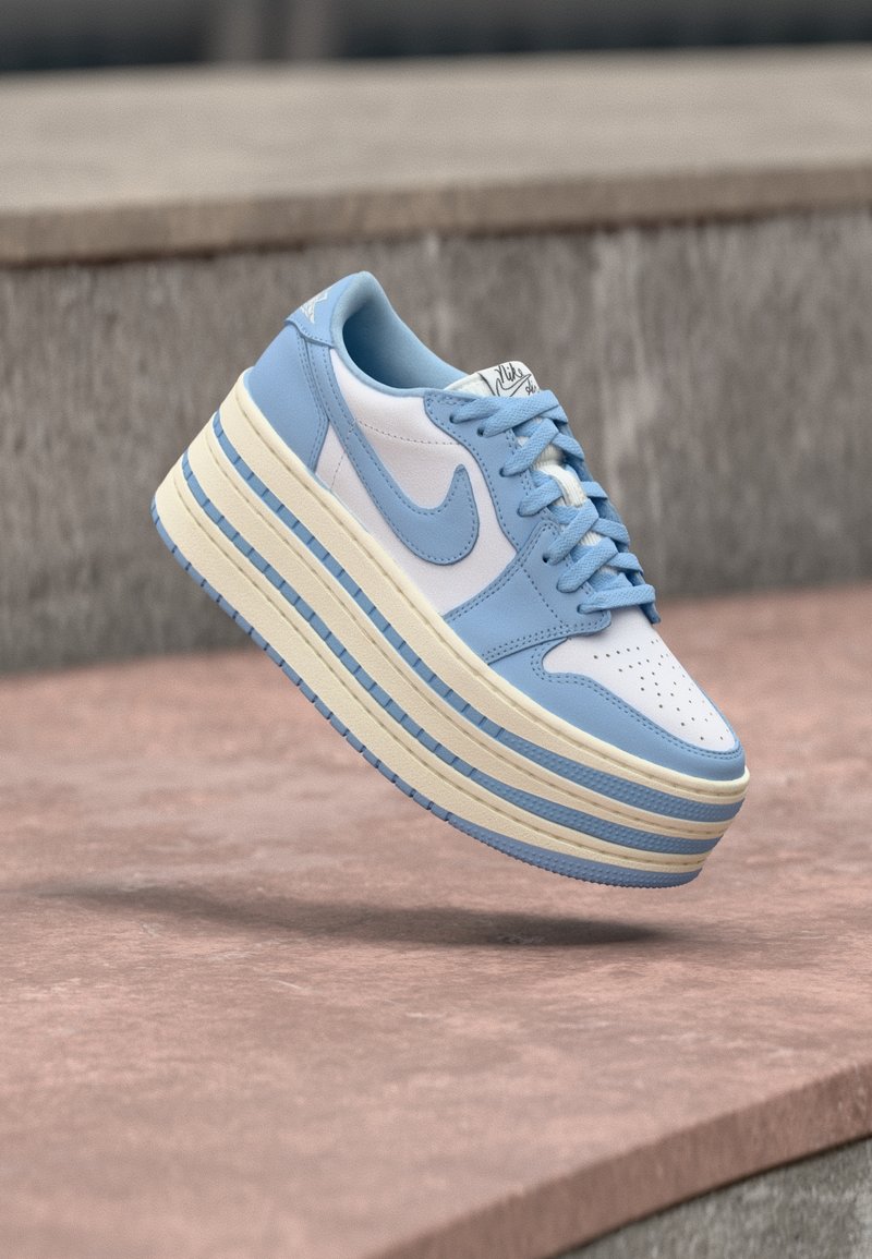 Baskets Nike blanches et bleu clair avec une semelle superposée surdimensionnée, flottant au-dessus d'une surface de pierre rosée et d'un fond en pierre grise.