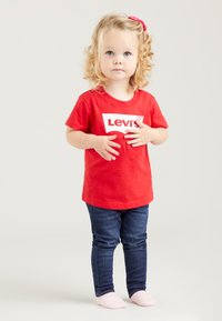 Rotes Baumwoll-T-Shirt mit weißem Levi's-Logo, kombiniert mit dunkelblauen Denim-Jeans. Das Kind hat lockiges Haar und trägt weiche rosa Socken.
