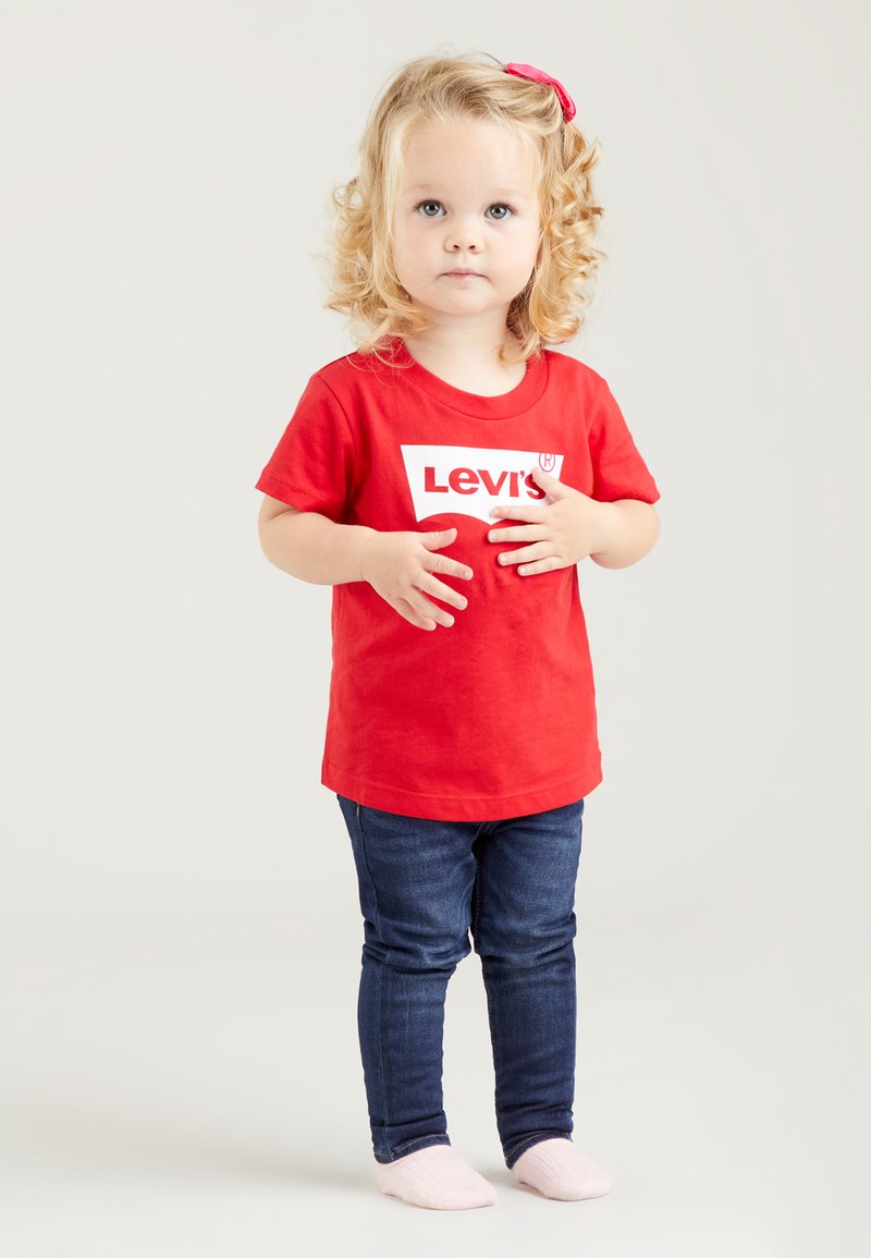 Rotes Baumwoll-T-Shirt mit weißem Levi's-Logo, kombiniert mit dunkelblauen Denim-Jeans. Das Kind hat lockiges Haar und trägt weiche rosa Socken.