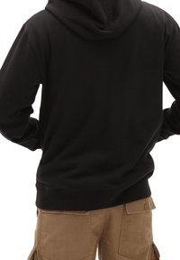 Person trägt eine schwarze Kapuzenjacke und braune Cargohosen, von hinten gesehen, Hände auf den Hüften ruhend vor weißem Hintergrund.