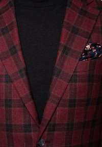 Czerwono-czarny żakiet w kratę z czarnym golfem pod spodem. Posiada niebieską kwiatową chusteczkę w kieszeni, podkreślającą finezyjne detale i teksturę.