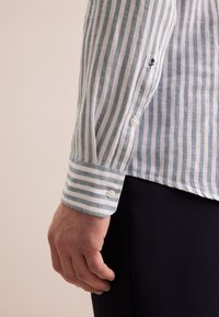 Gestreepte shirtmouw in lichtblauw en wit, gemaakt van ademende stof, met knoopsluitingen en een subtiele geborduurde detail op de manchet.